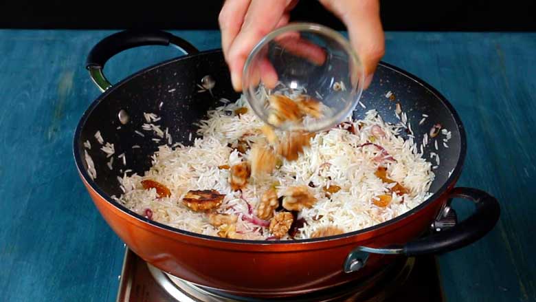 A hand is seen tipping roasted California walnuts from a small glass bowl into rice being cooked in an orange non stick pan on an electric stove on a blue wooden counter top - Kerala Ghee Rice with California Walnuts Recipe